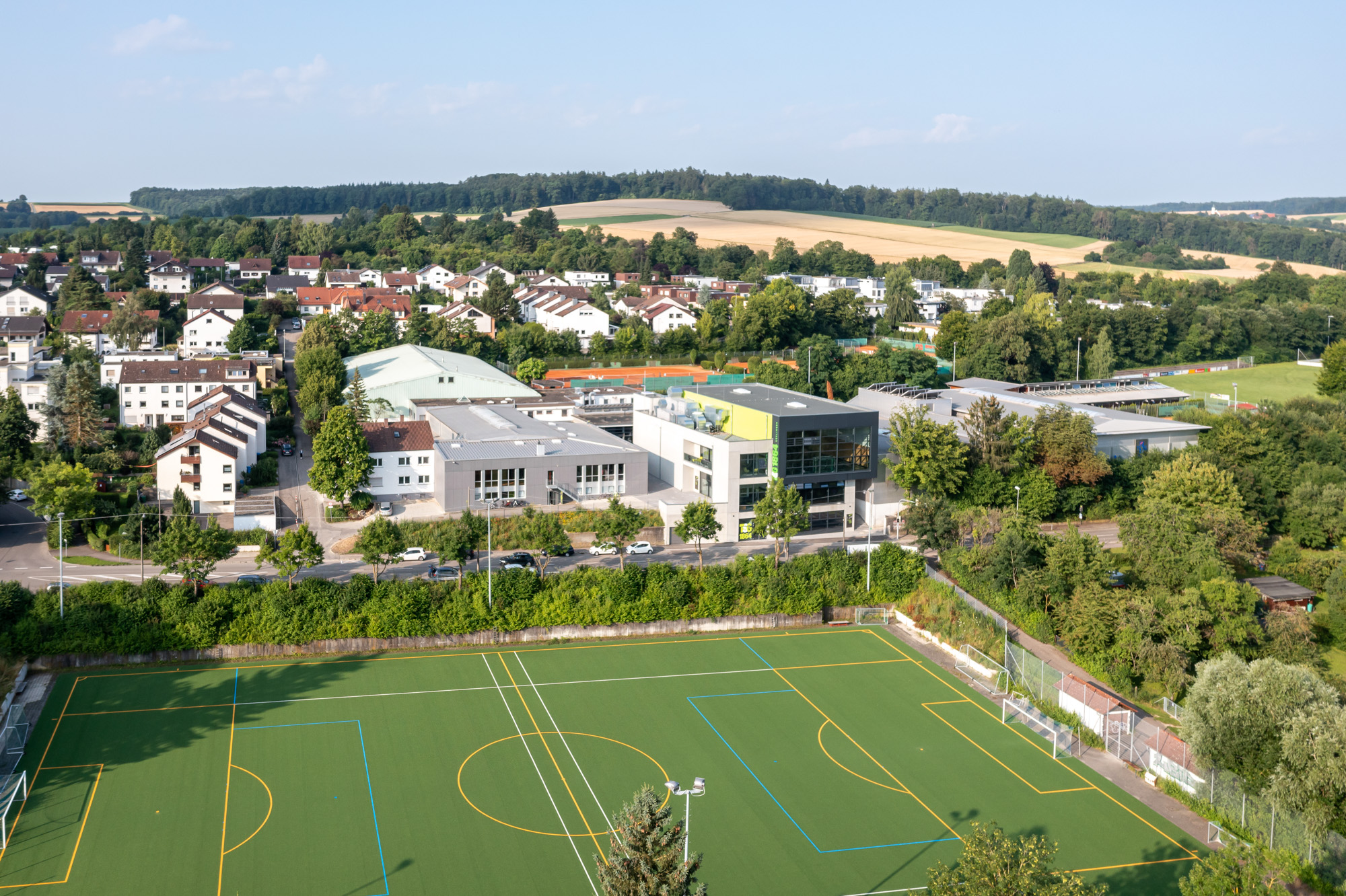Aerial view of a sports complex featuring a green artificial turf soccer field with yellow and blue markings, modern buildings, and a residential neighborhood surrounded by trees and rolling hills under a clear sky.