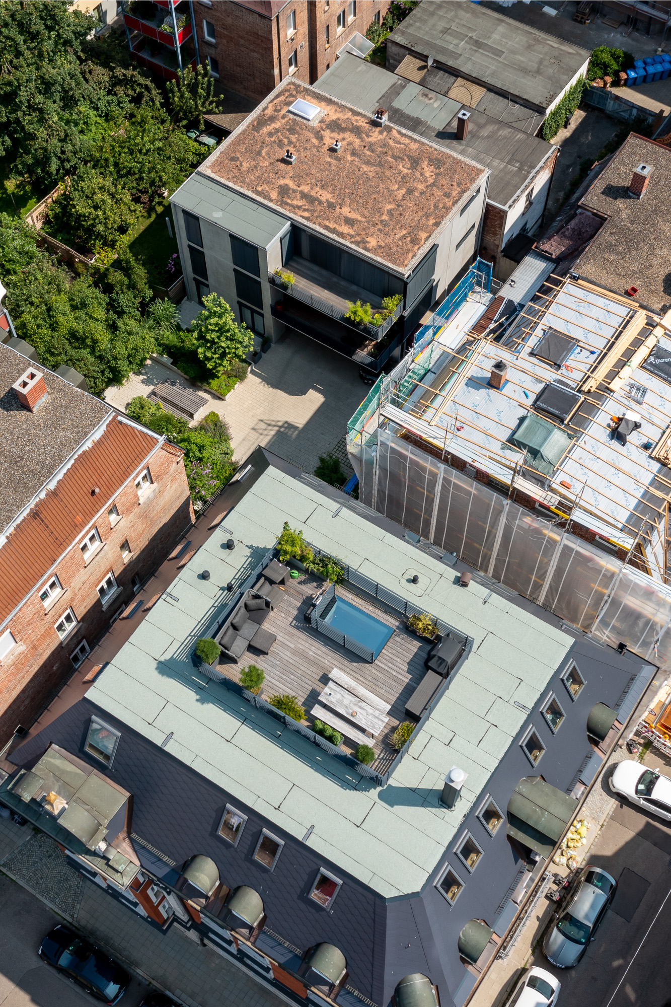 Aerial view of urban rooftops with a central rooftop terrace featuring outdoor seating, a table, potted plants, and a small pool.