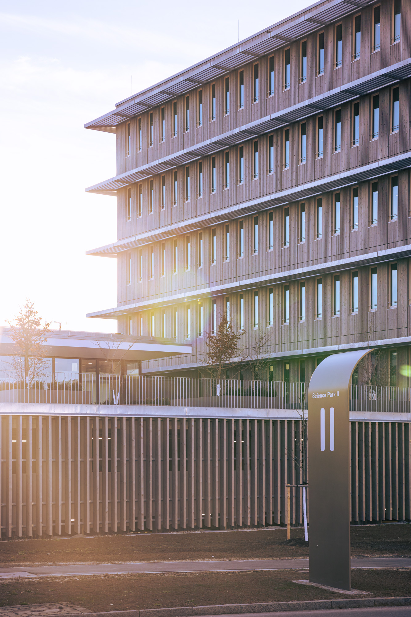 Modern multi-story building with wooden paneling and rows of windows, labeled Science Park II, during sunset with sunlight flare.