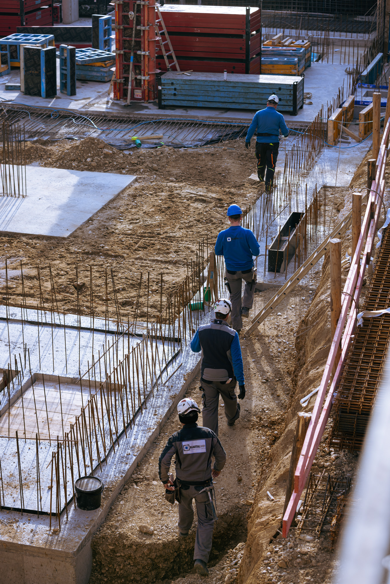 Four construction workers in safety helmets walking along a dirt path at a construction site with metal rod reinforcements and stacked materials.