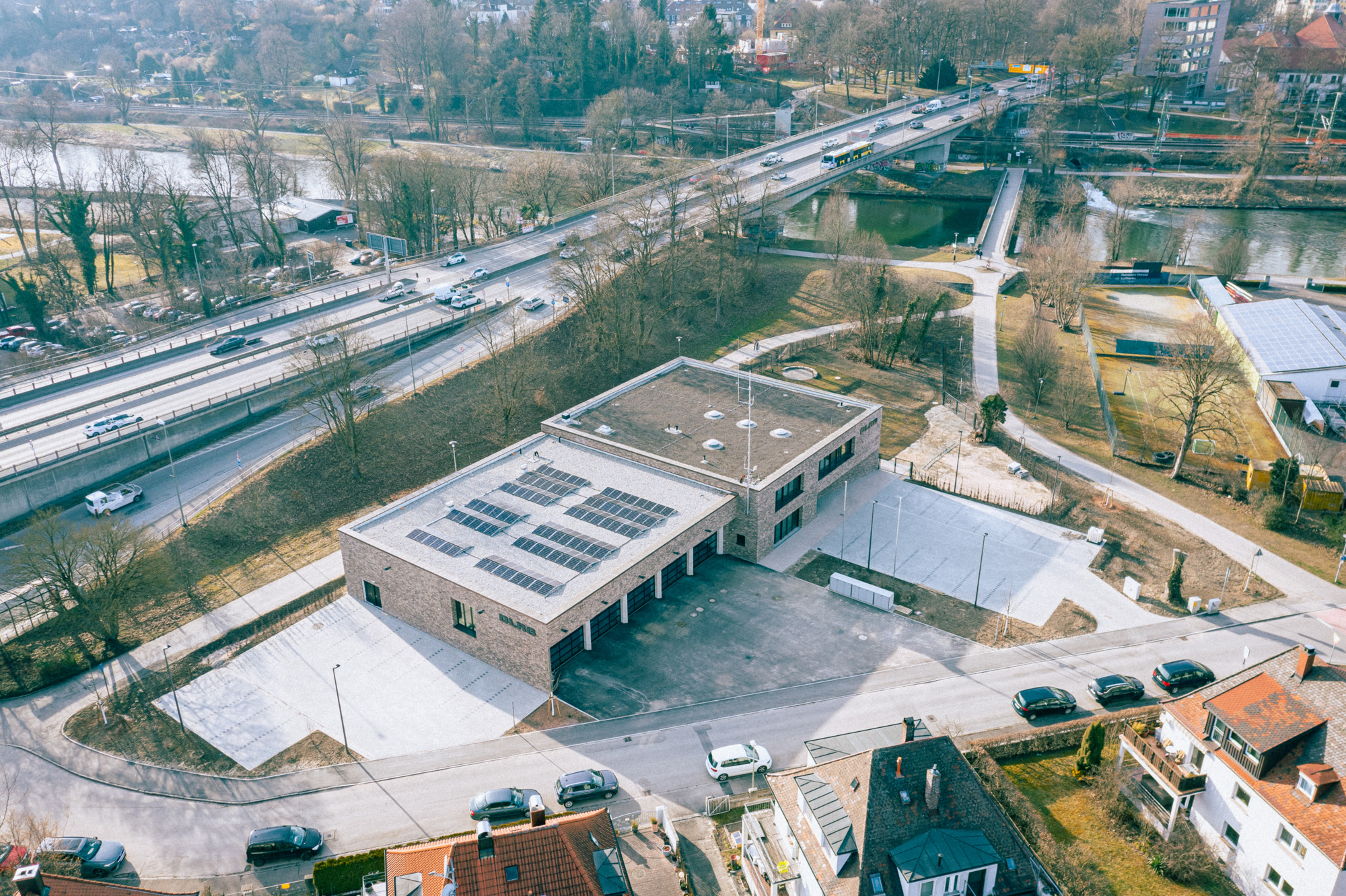Aerial view of a modern brick building with solar panels on the roof, surrounded by roads, parked cars, and a nearby river with bridges.