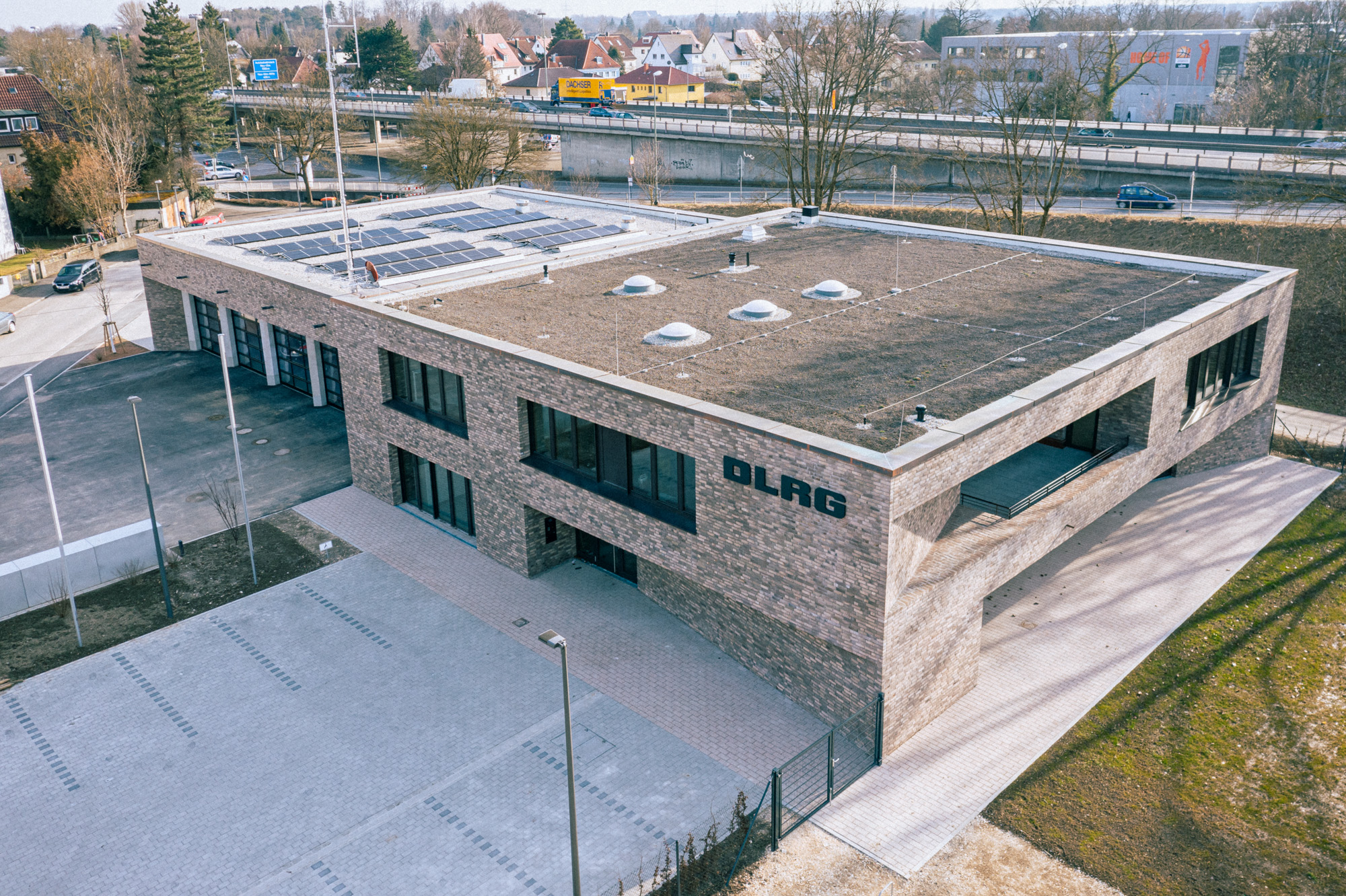 Modern two-story brick building with solar panels on the roof and large windows, labeled DLRG, with an empty paved parking area in front.