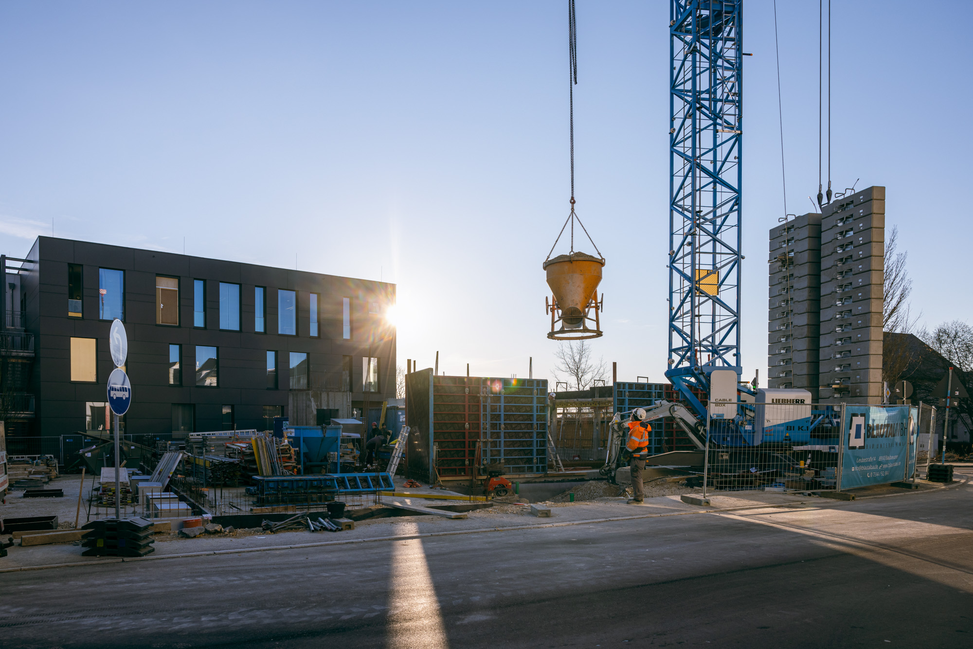 Construction site with a crane lifting a concrete bucket next to a modern building at sunset.