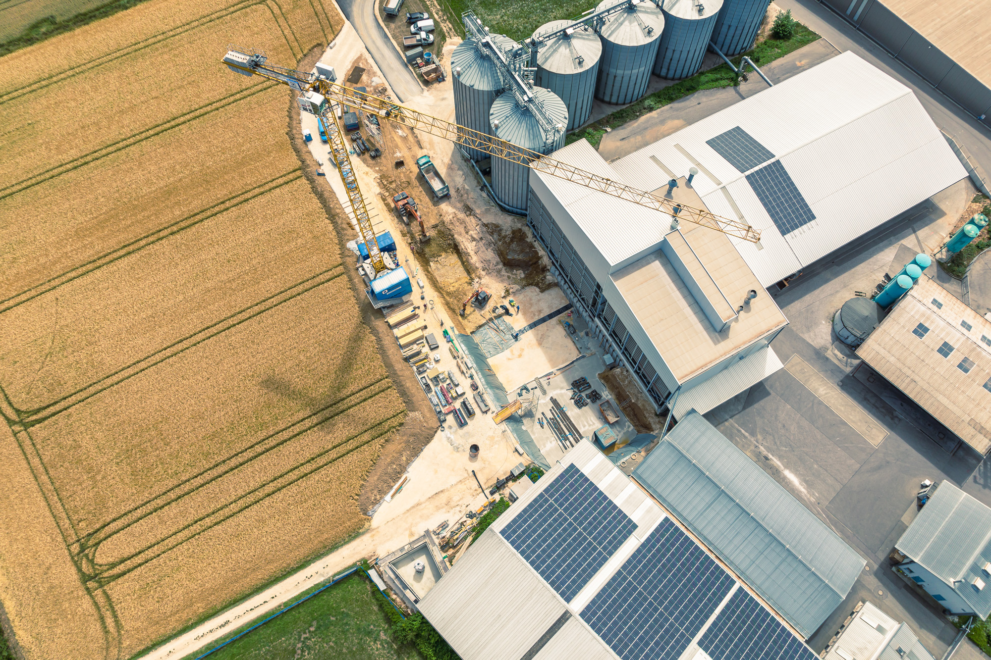 Aerial view of an industrial facility with buildings featuring solar panels, adjacent construction site, and surrounding farmland.