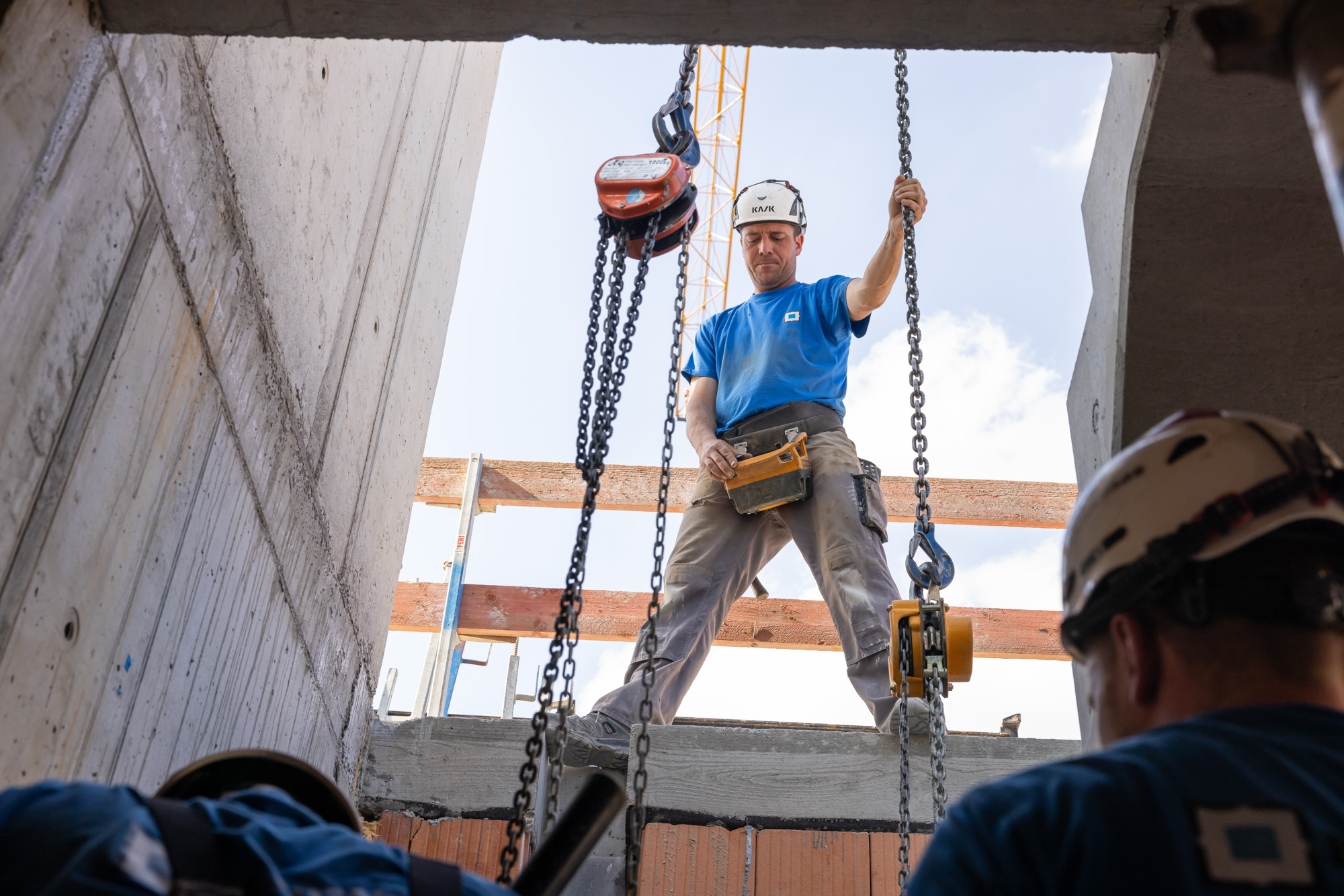 Construction worker in a blue shirt and helmet holding chains and operating hoist equipment at building site opening above two other workers.