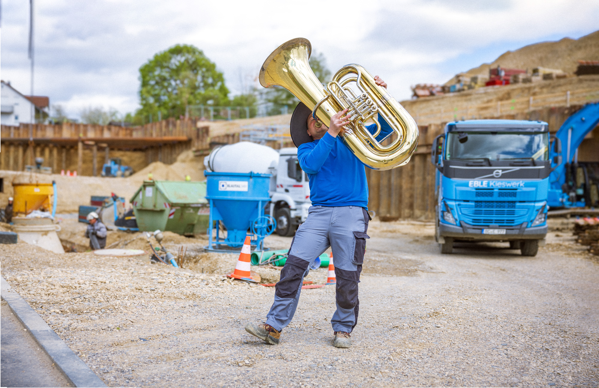 Person wearing a blue shirt and gray pants playing a large brass tuba at a construction site with trucks and equipment in the background.