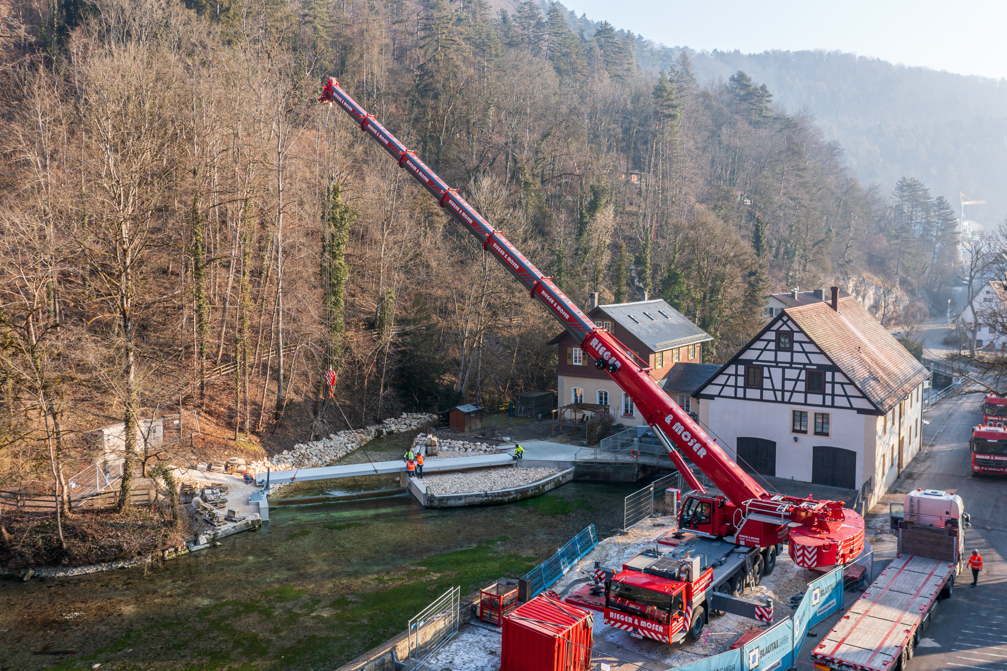 Large red crane lifting a long steel beam over a river near traditional houses and forested hills.