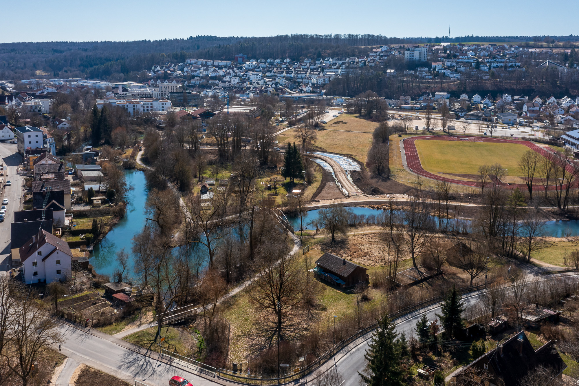 Aerial view of a small town with a winding blue river, residential houses, a running track, and wooded hills in the background.