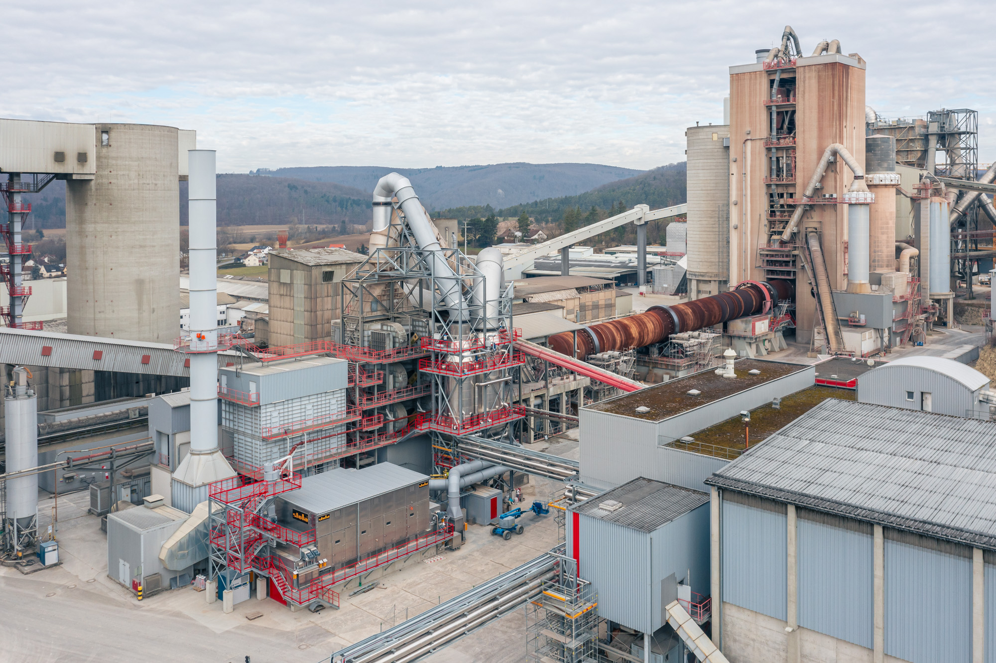 Industrial facility with multiple large buildings, ventilation pipes, and a rusted rotary kiln against a background of forested hills and cloudy sky.