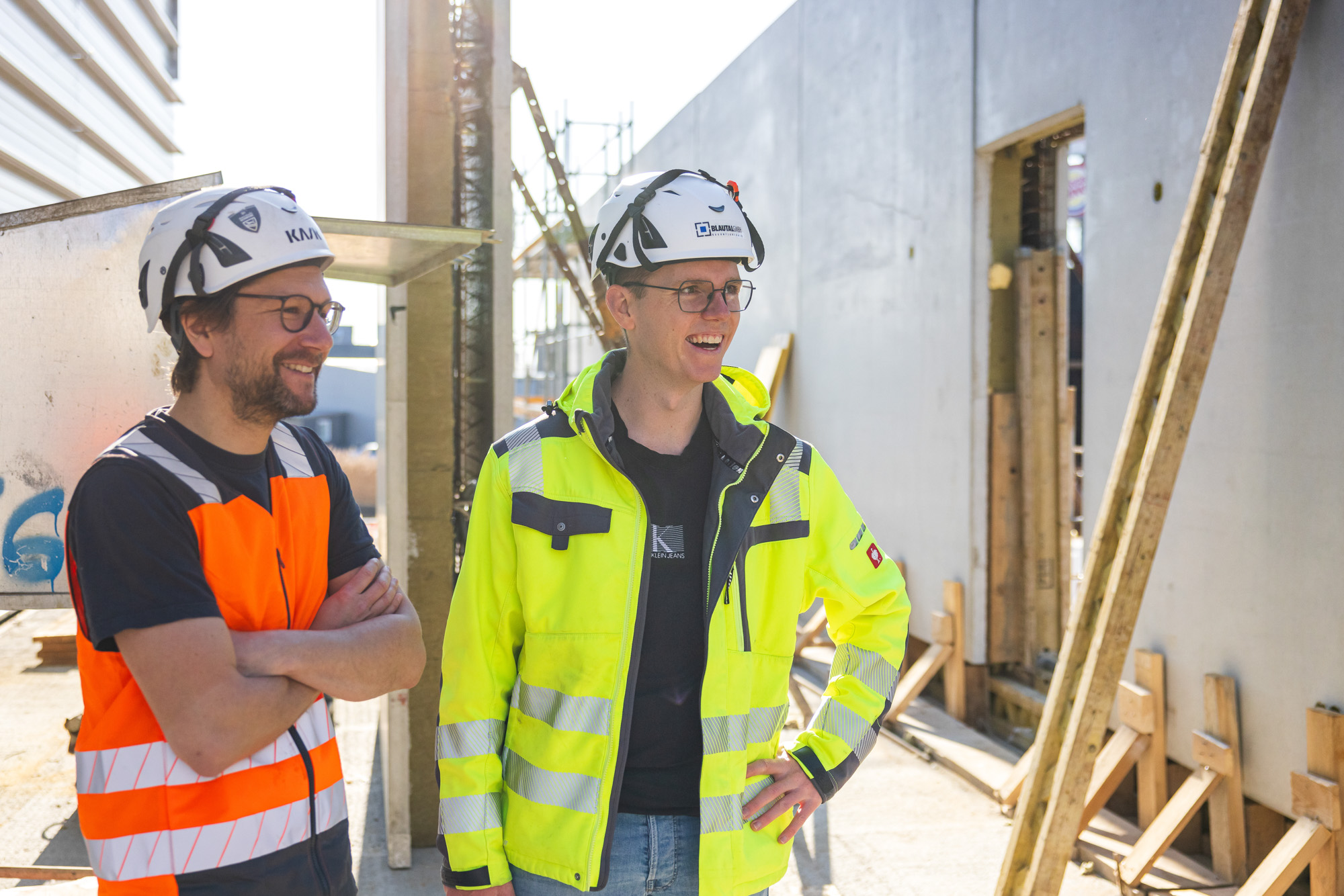 Two construction workers wearing helmets and safety vests stand and smile at a building site with scaffolding and construction materials.