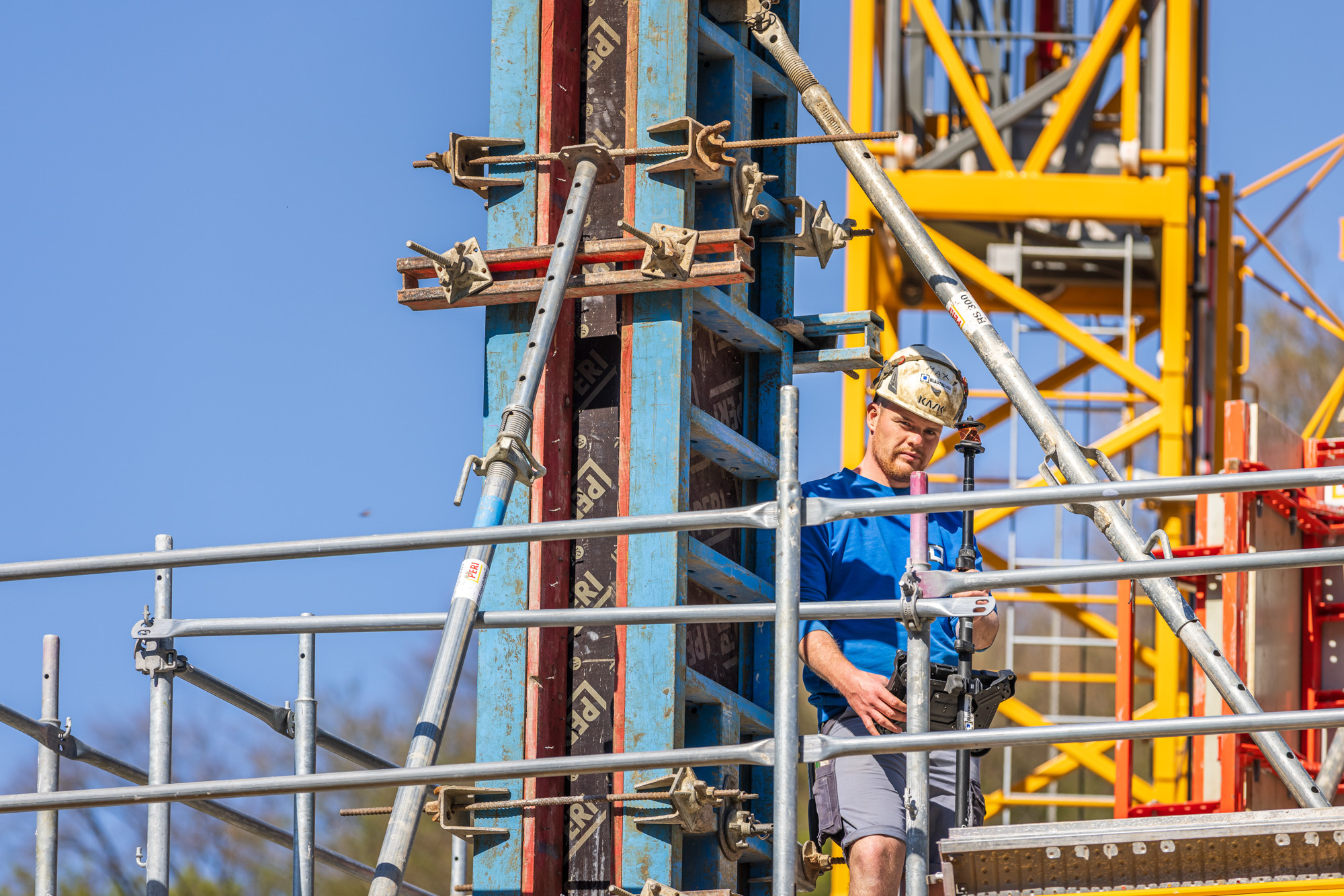Construction worker wearing a helmet and blue shirt working on scaffolding near a blue and yellow building structure against a clear sky.