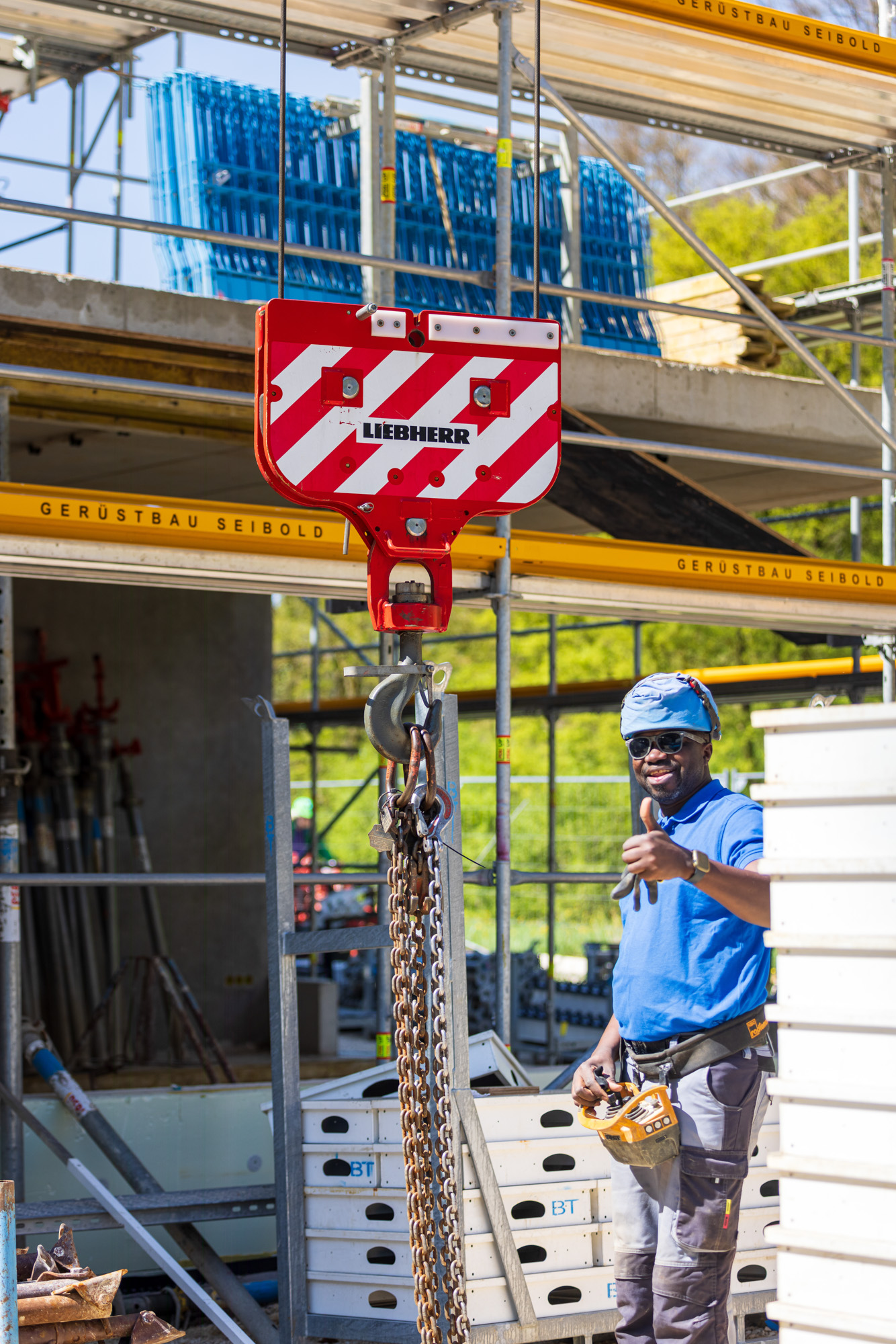 Construction worker in blue shirt and helmet giving thumbs up next to a red and white Liebherr crane hook on a building site.