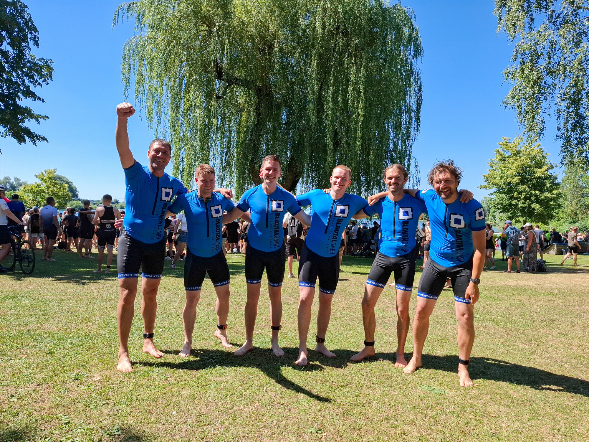 Six men in matching blue and black triathlon suits standing barefoot on grass with arms around each other under a large tree on a sunny day.