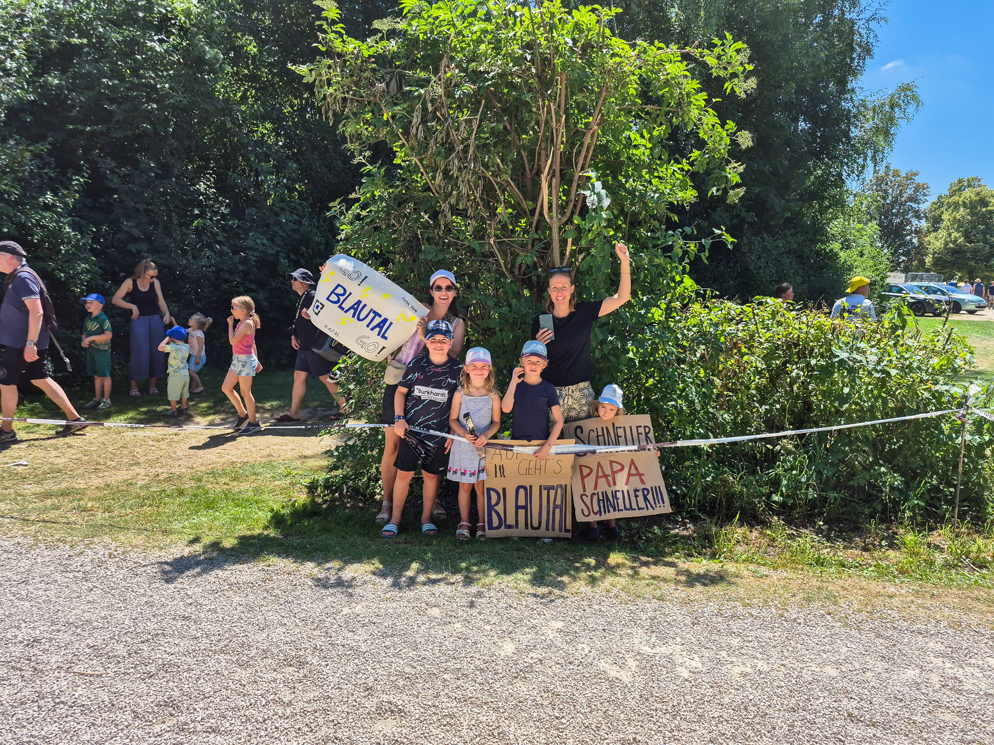 Group of adults and children outdoors holding signs with supportive messages for Blautal, cheering at a sunny event.