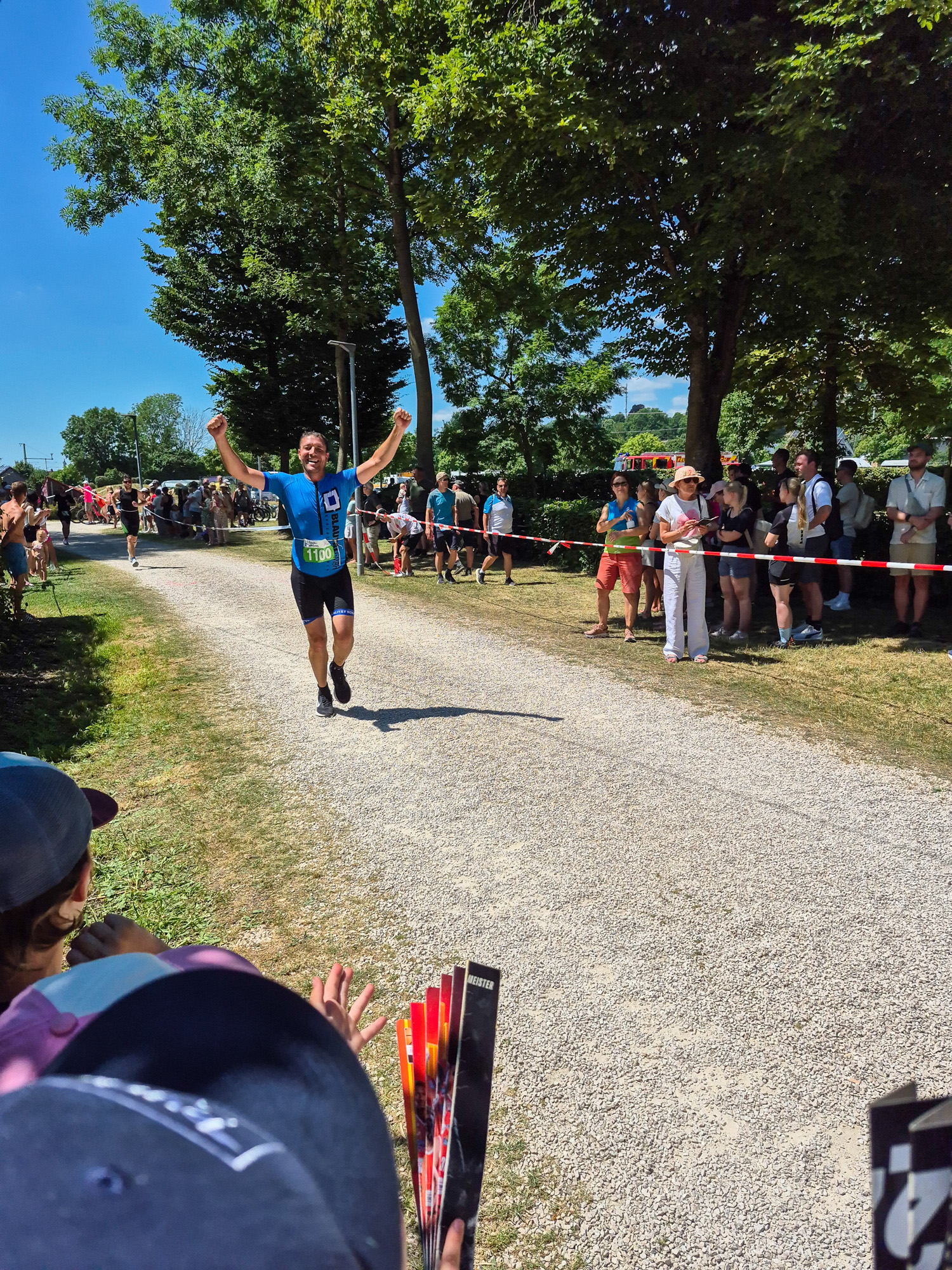 Smiling male runner in blue shirt raising arms near finish line of outdoor race with spectators on either side of gravel path.