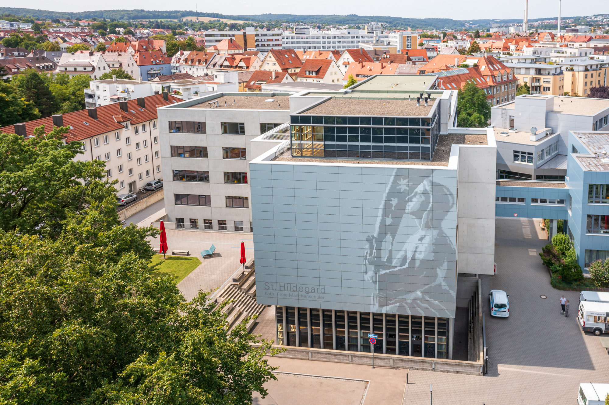 Modern school building with a large grayscale portrait mural and a courtyard with red umbrellas.