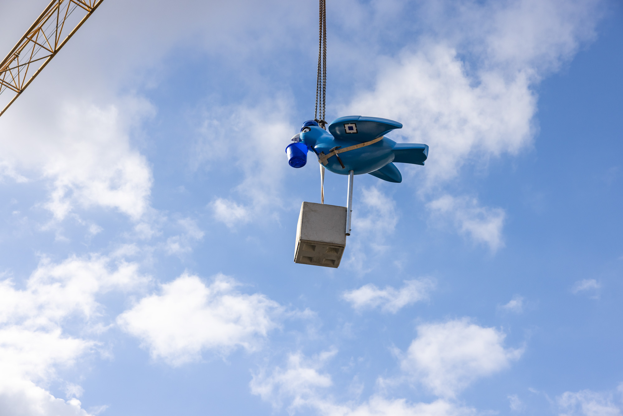 Blue crane hook shaped like a cartoon bird carrying a concrete block against a partly cloudy sky.