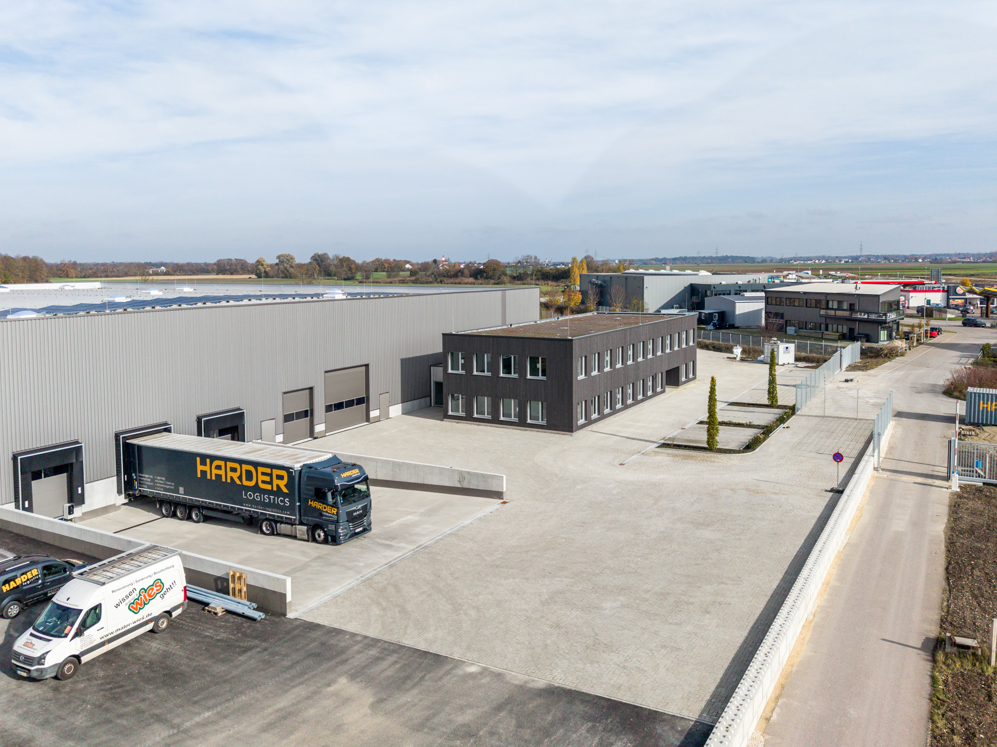 Industrial warehouse complex with a black HARDER Logistics truck parked at a loading dock and a modern two-story office building nearby under a cloudy sky.