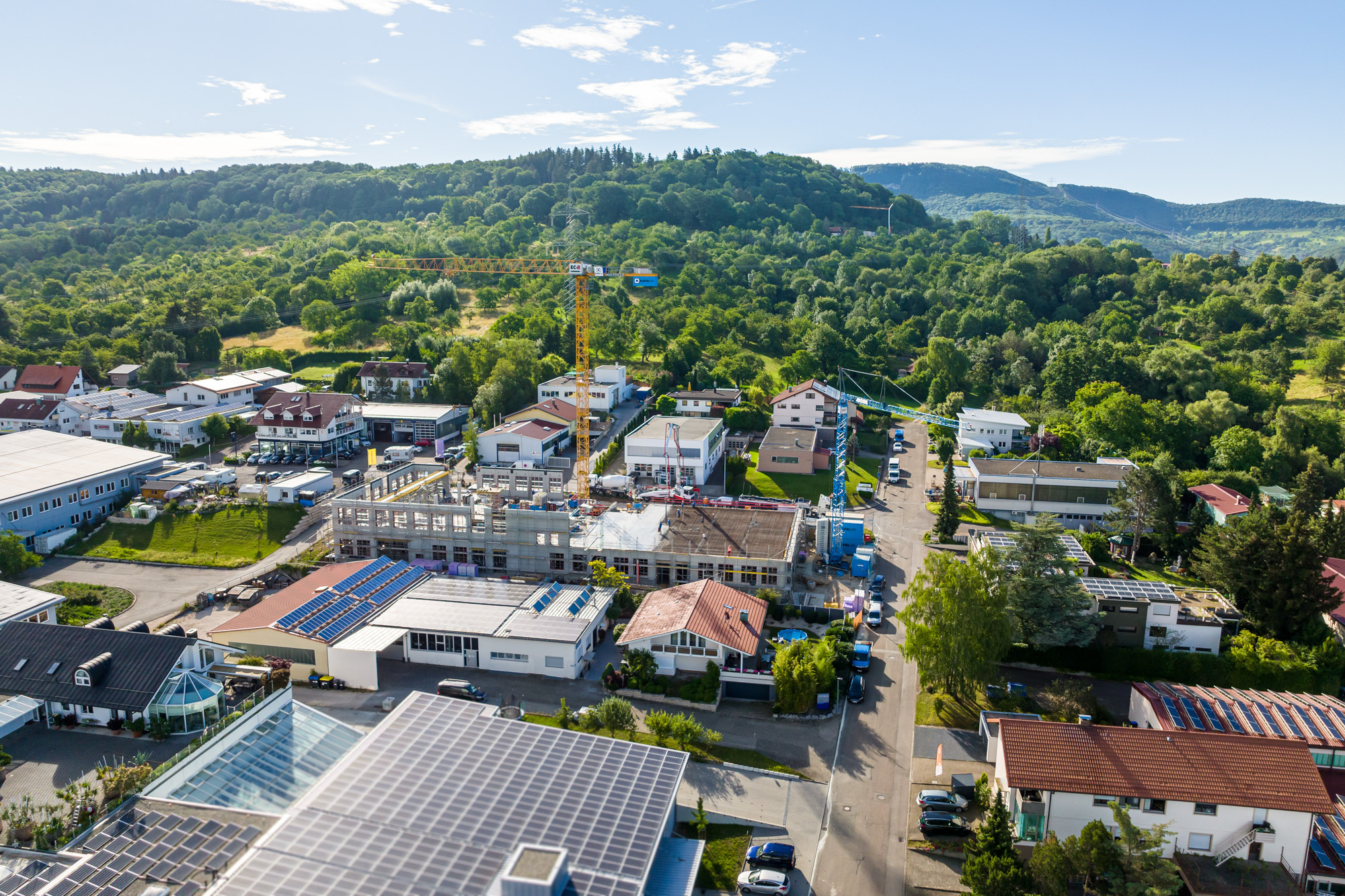 Aerial view of a suburban area with a construction site featuring cranes, surrounded by houses with solar panels and green hills in the background.