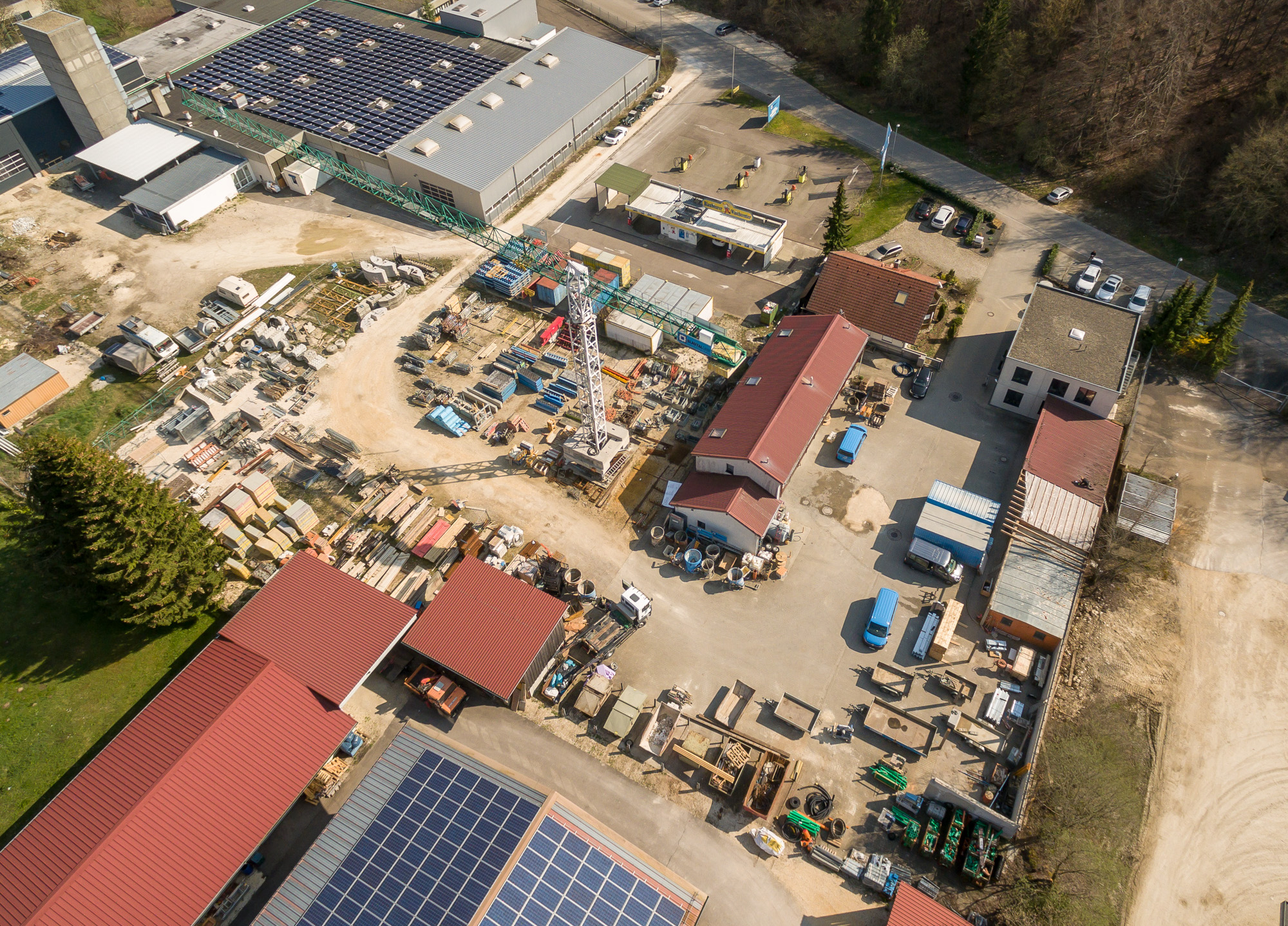 Aerial view of an industrial yard with storage buildings, construction materials, a crane, and solar panels on rooftops.