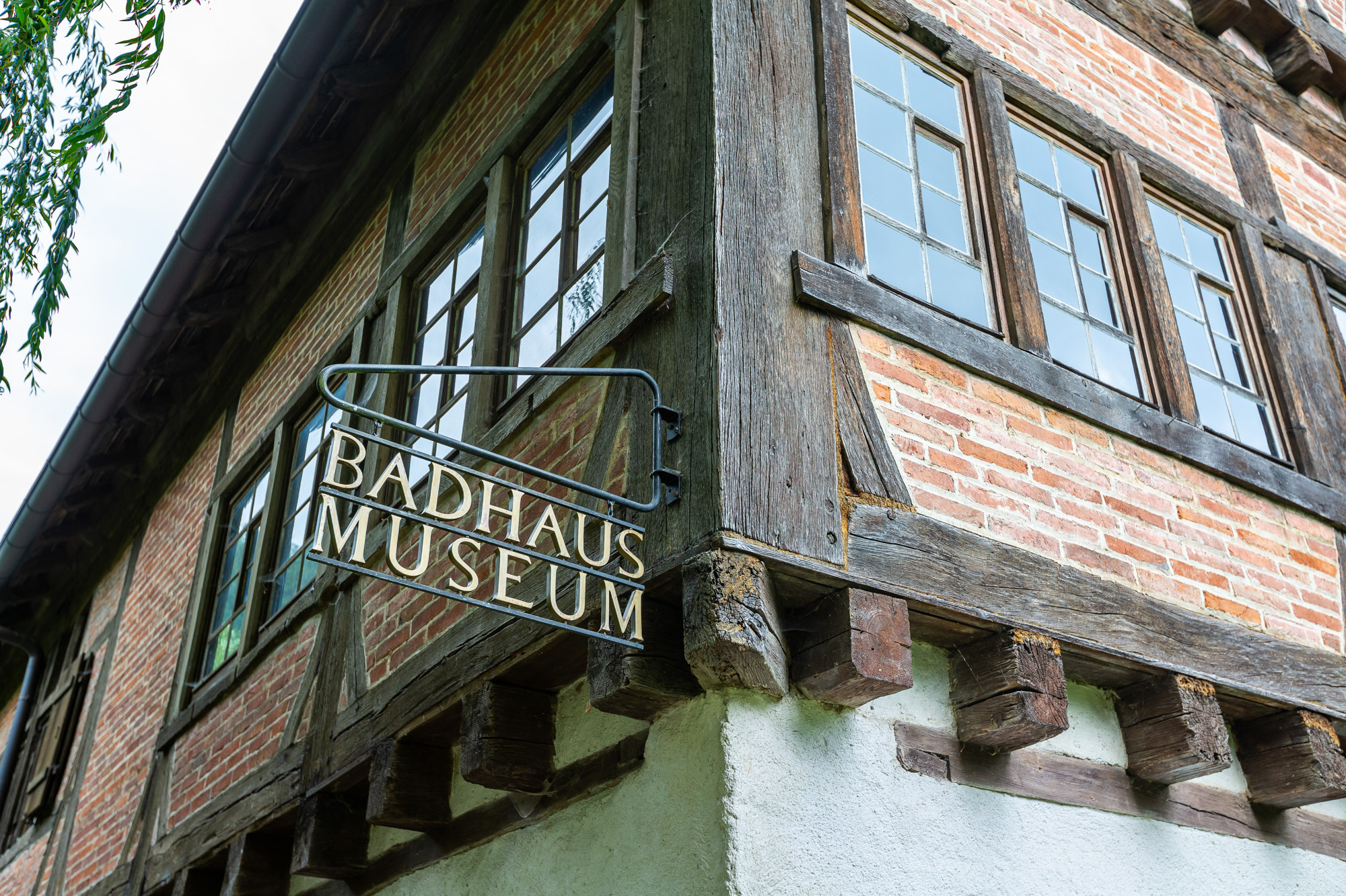 Corner of a historic half-timbered brick building with a metal sign reading 'BADHAUS MUSEUM'.