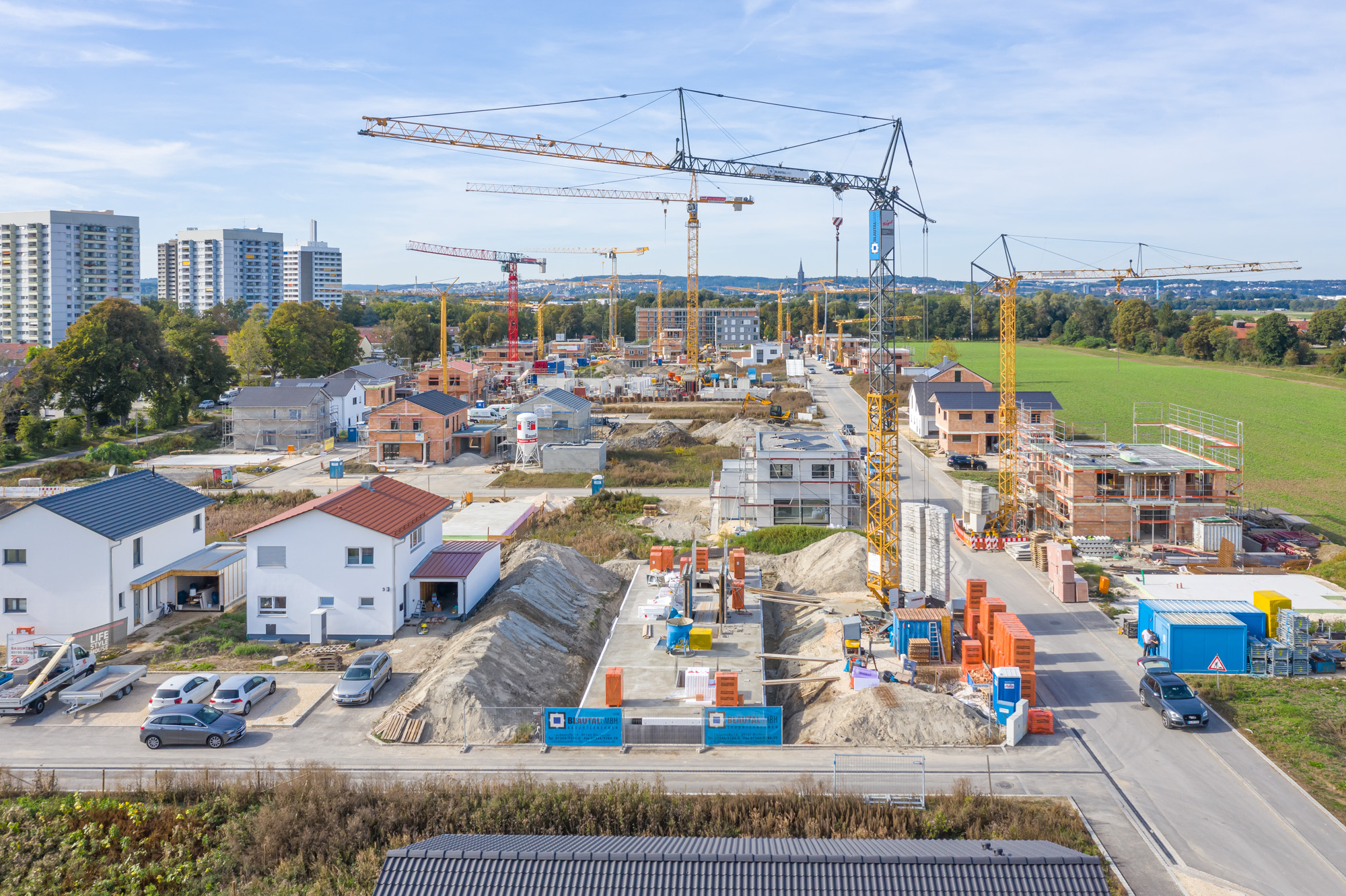 Aerial view of a residential construction site with multiple cranes, partially built houses, and parked cars under a blue sky.