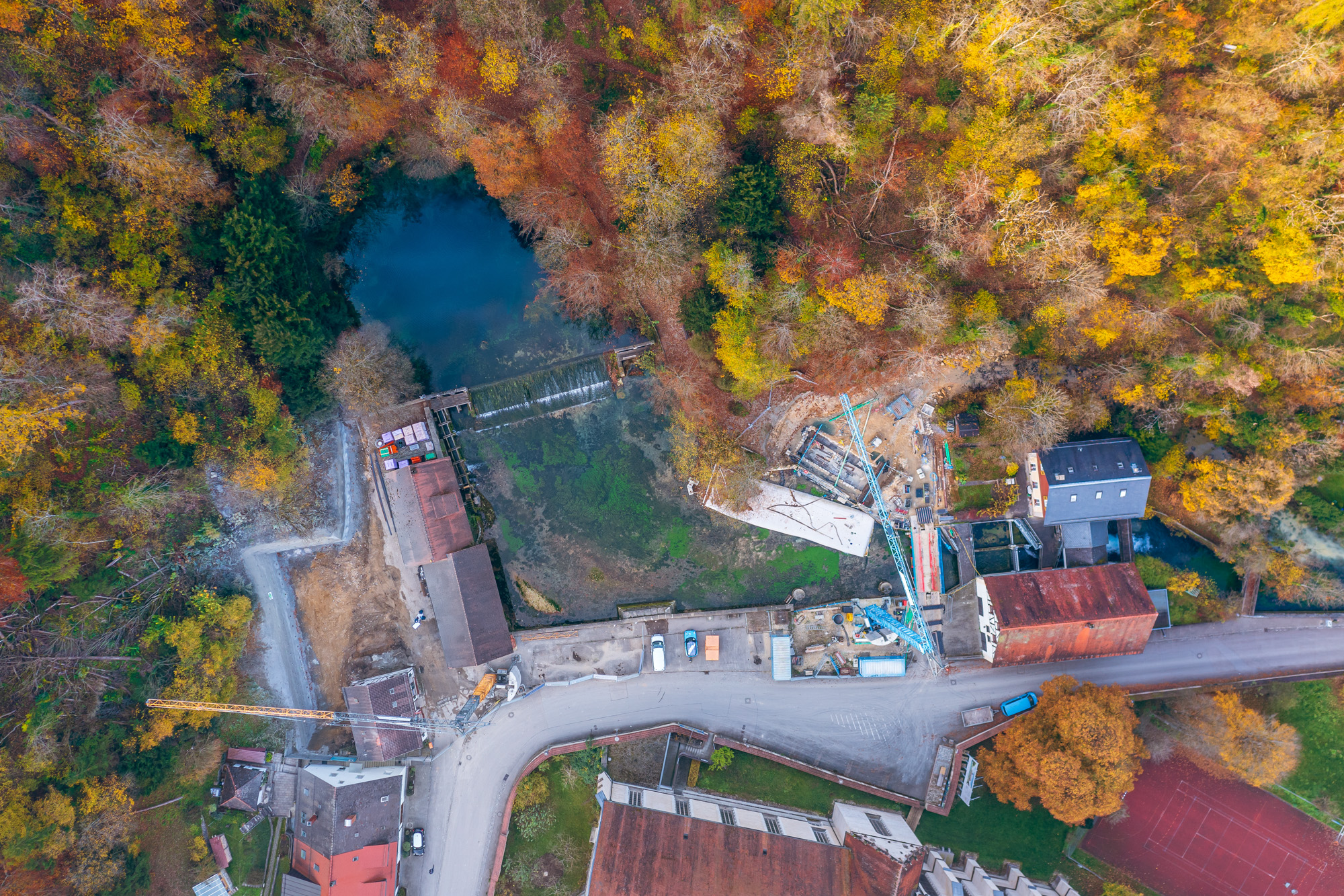 Aerial view of a small river or pond surrounded by autumn-colored trees with buildings, construction cranes, and a curved road nearby.
