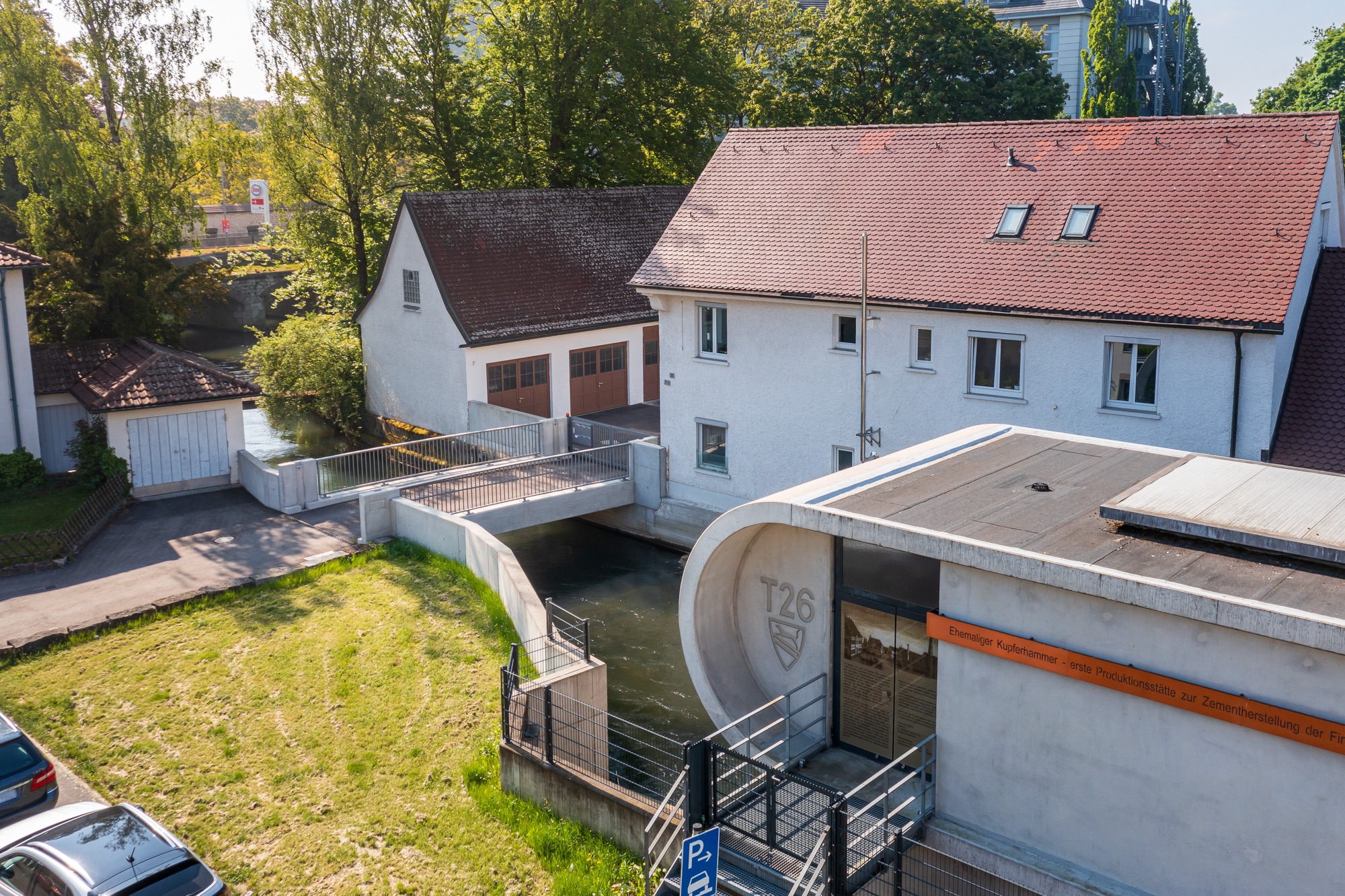 A river flowing under a pedestrian bridge next to white buildings with red tiled roofs surrounded by green trees and parked cars.