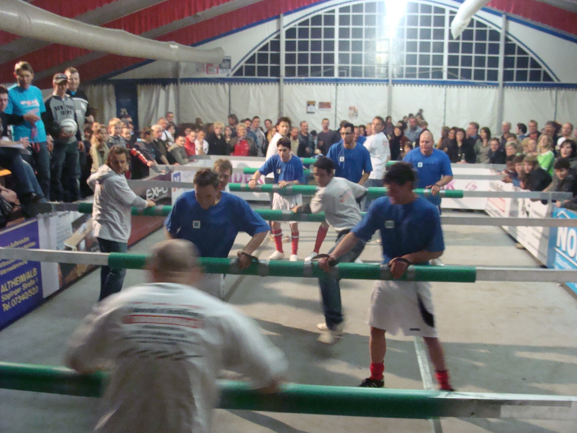 Indoor human foosball game with two teams in blue and white shirts surrounded by a crowd of spectators.