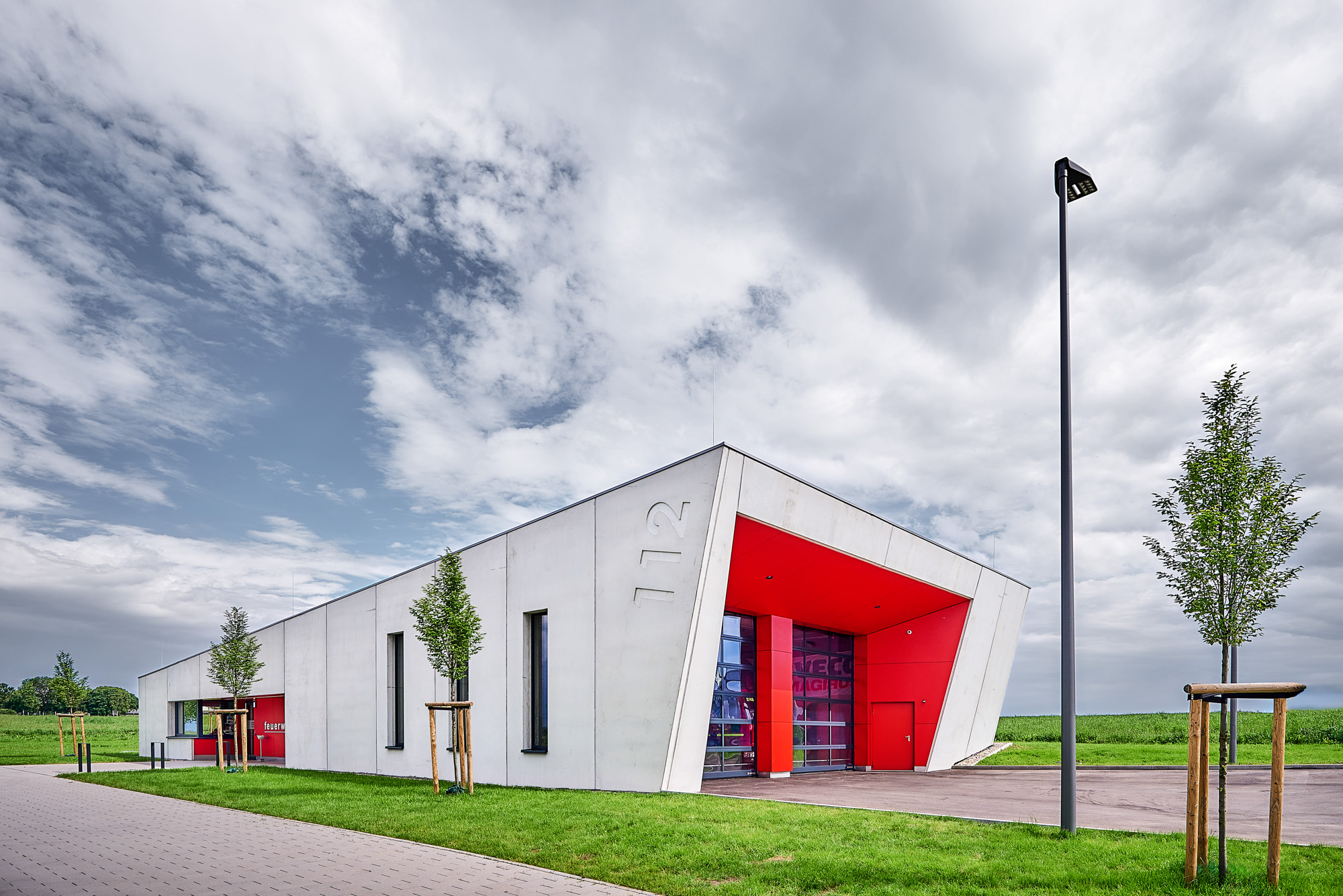 Modern fire station building with angled white walls, red entrance, and number 112 on the side, surrounded by green grass and trees under a partly cloudy sky.