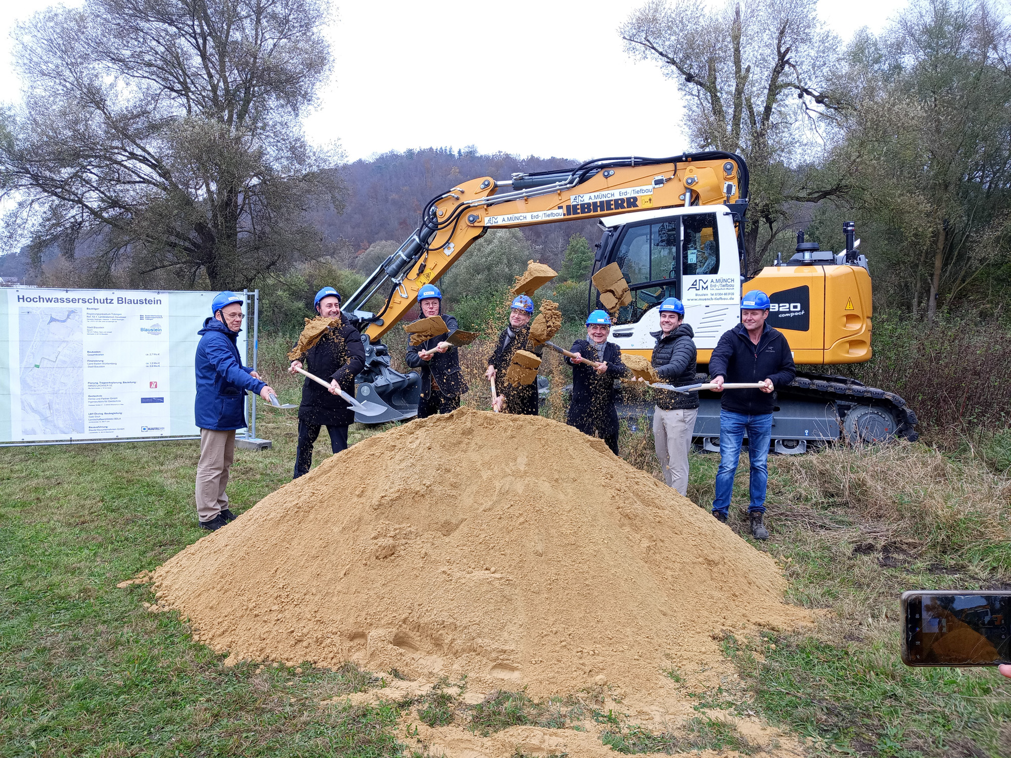 Seven people wearing blue safety helmets and holding shovels throw dirt in front of a construction excavator and a large pile of sand with a project sign in the background.