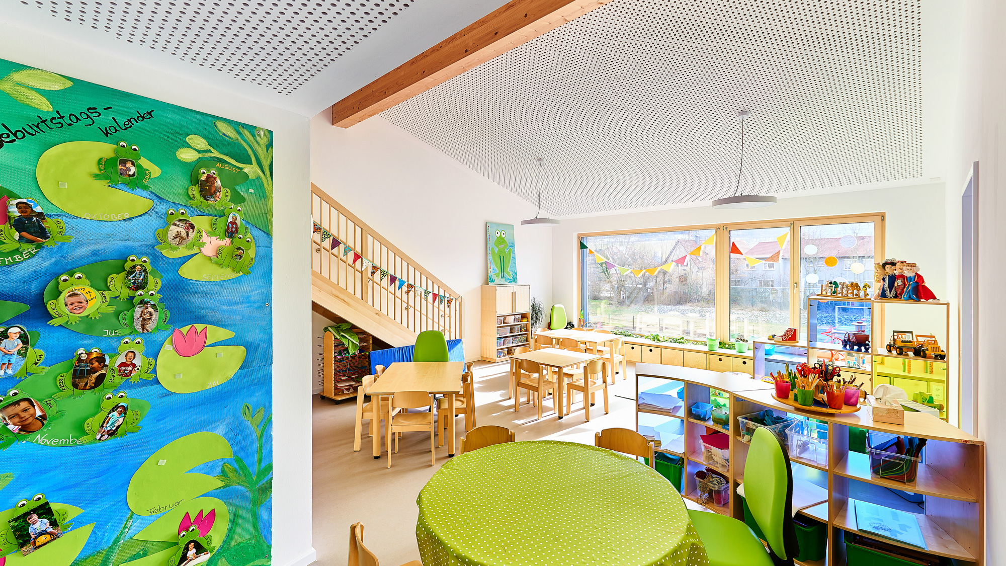 Bright kindergarten classroom with small wooden tables and chairs, colorful storage bins, a birthday photo board with frog cutouts, and large windows with outdoor view.