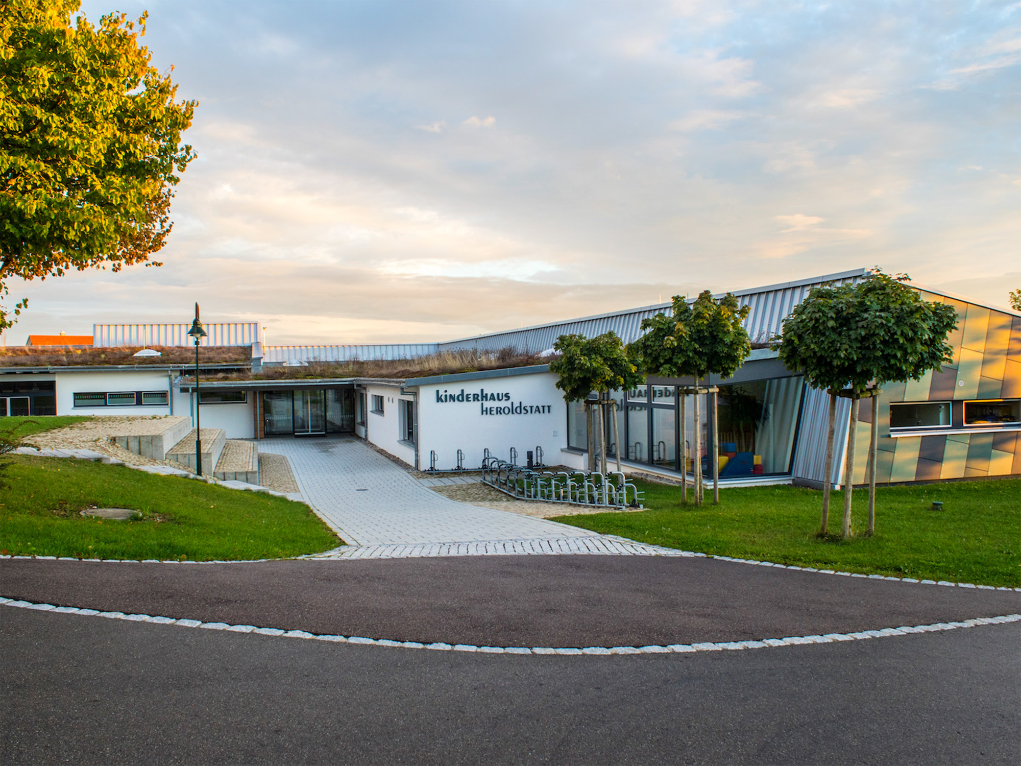 Modern building with a sloped roof and large windows, labeled Kinderhaus Heroldstatt, surrounded by green lawns and young trees under a cloudy sky.