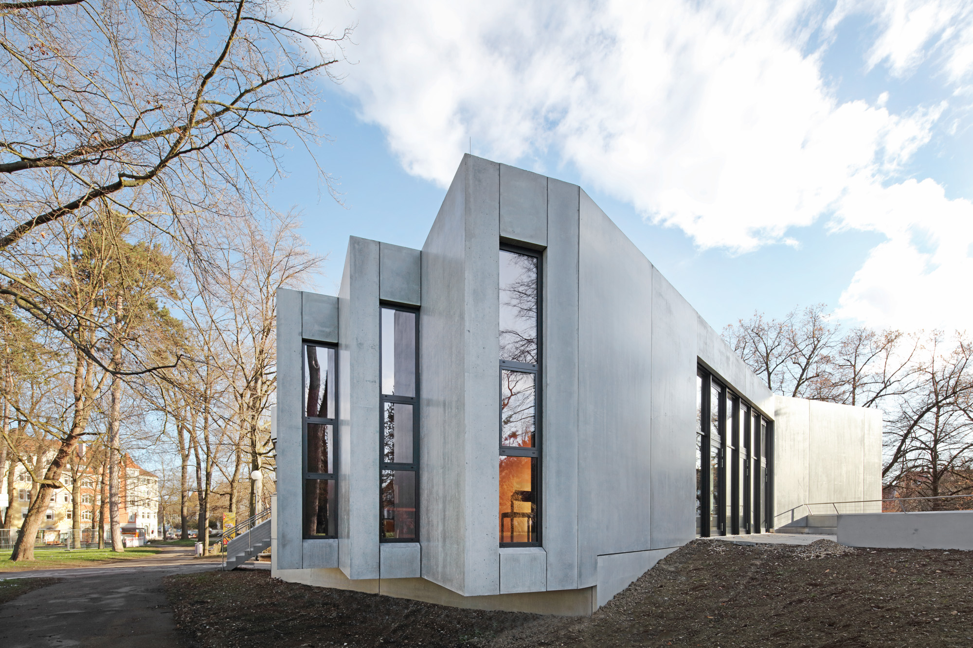 Modern concrete building with tall vertical windows surrounded by leafless trees under a partly cloudy sky.
