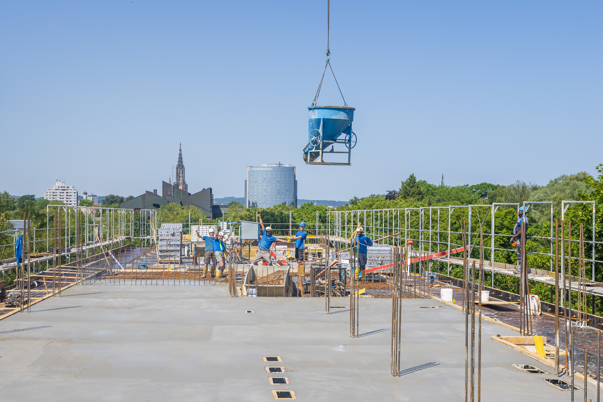 Construction workers on a building site with a suspended blue concrete bucket above them and city buildings and trees in the background under clear blue sky.