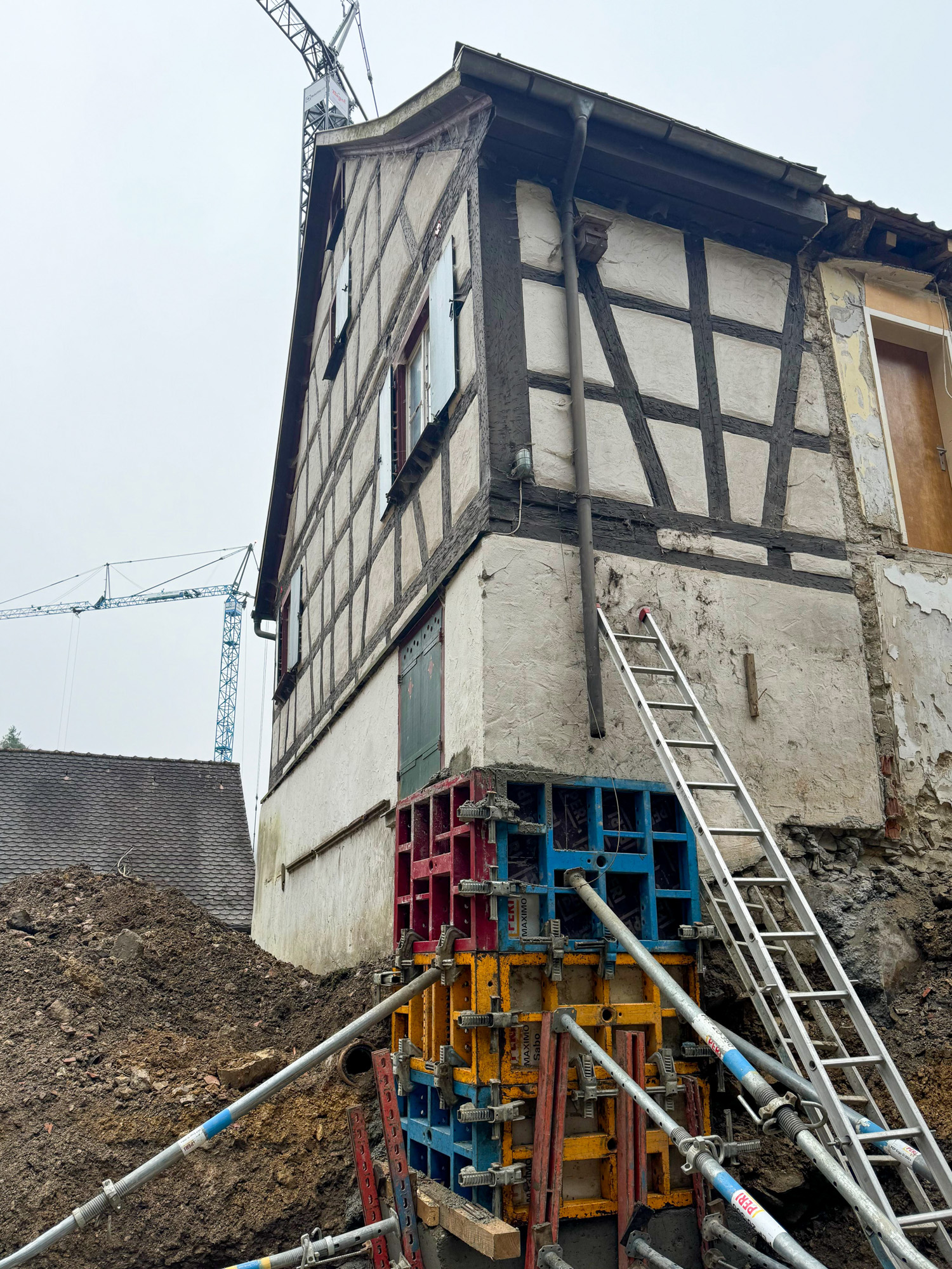 Half-timbered house under construction with colorful scaffolding formwork and metal support beams around the foundation, a tall ladder leaning against the wall, and cranes in the foggy background.