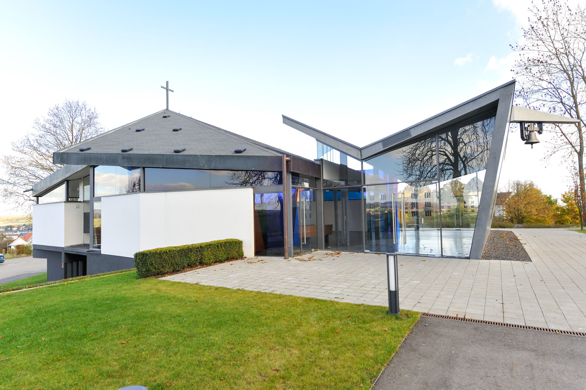 Modern church building with a cross on a gray sloped roof and glass walls reflecting surrounding trees.