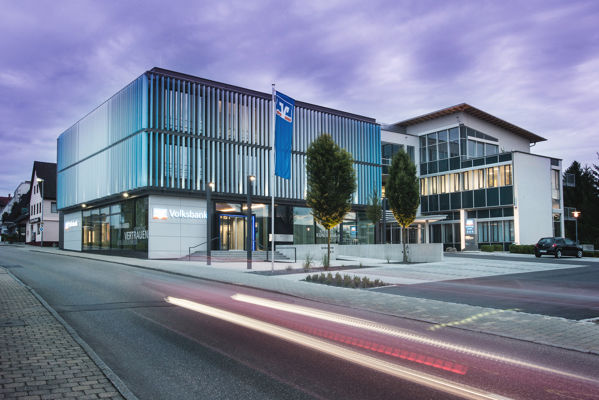 Modern Volksbank building with vertical metal slats exterior and illuminated signage at dusk, with light trails from passing cars.